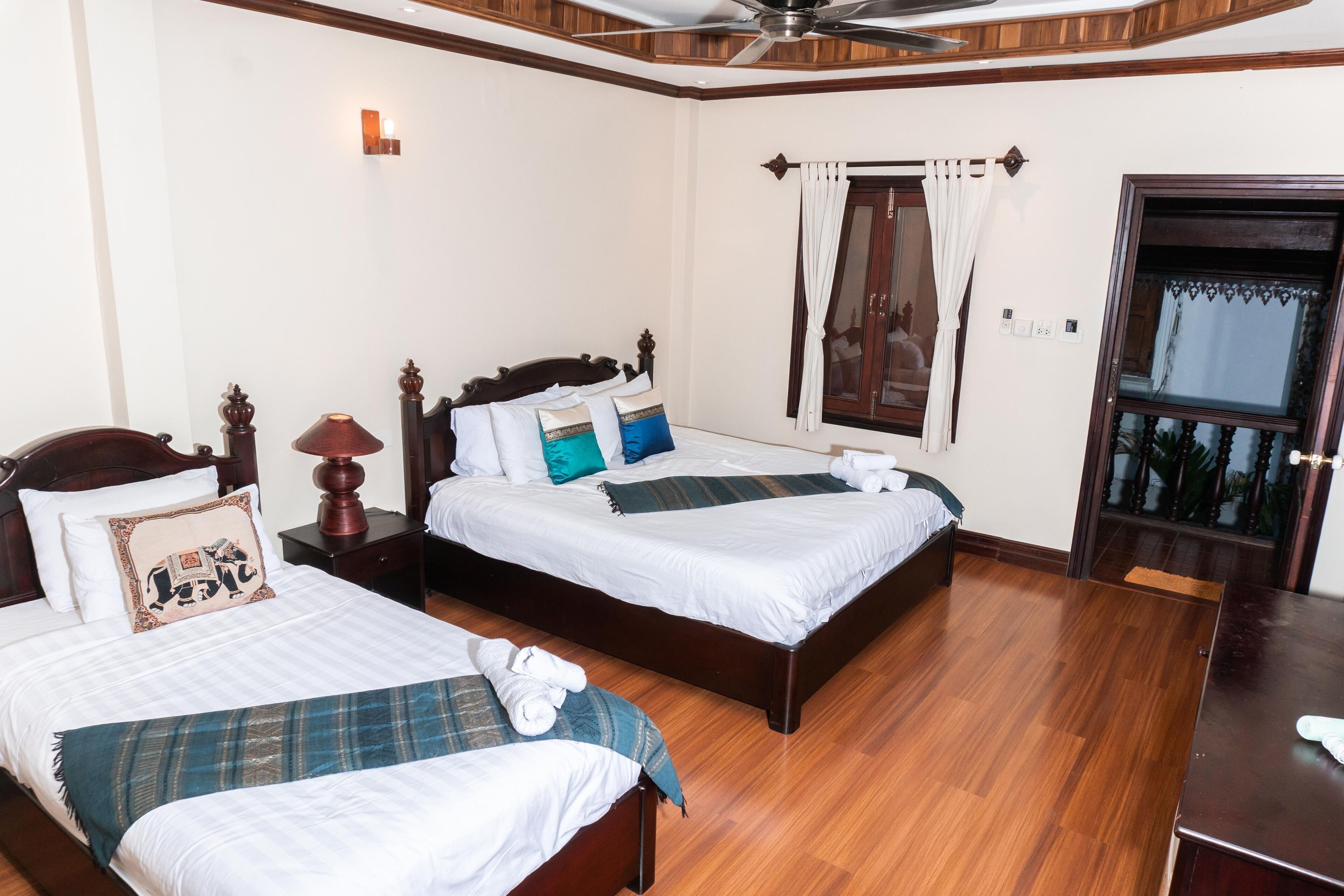 A hotel room with two beds, wooden flooring, a ceiling fan, and a window with curtains.