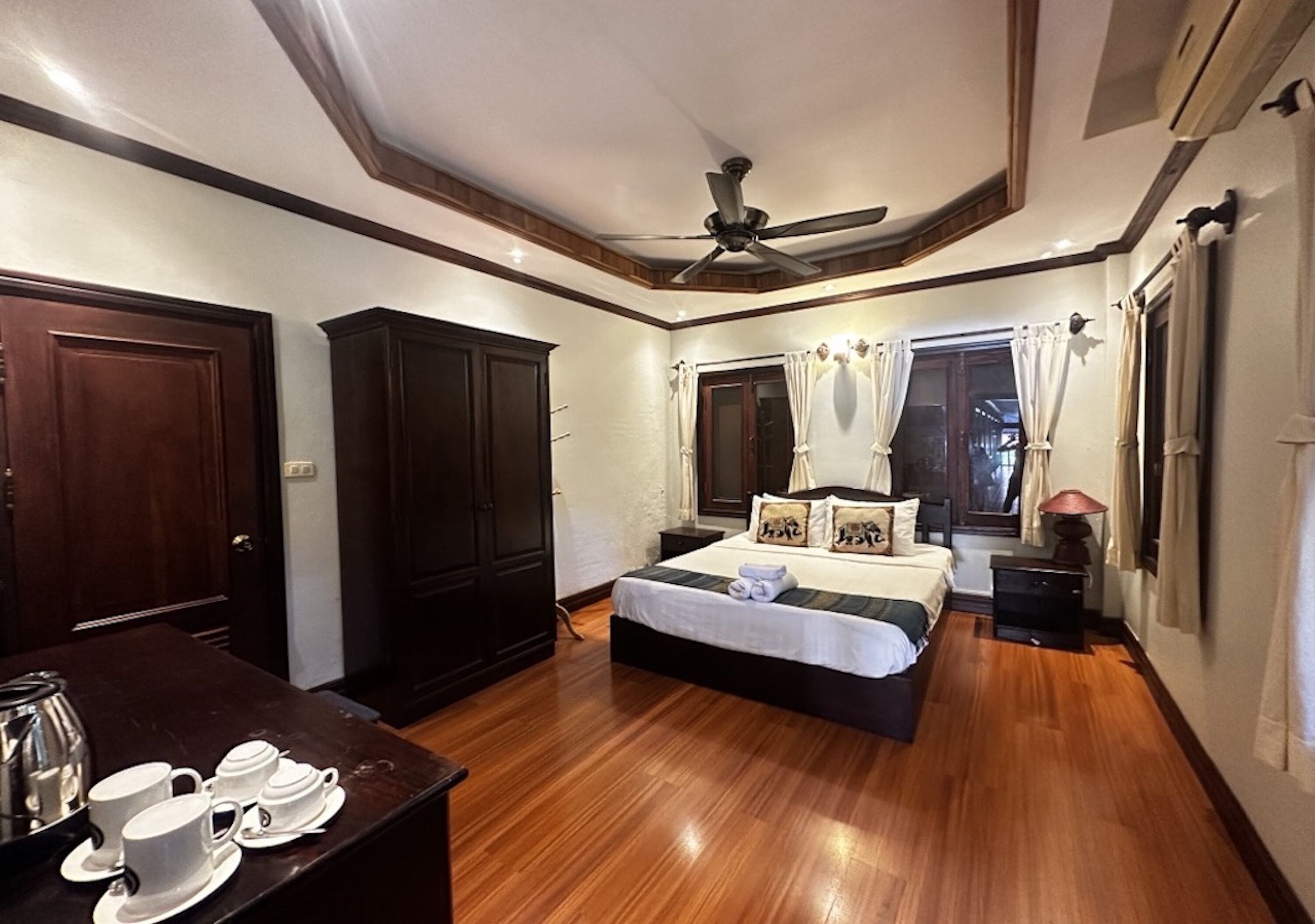 A bedroom with a bed, wooden floor, ceiling fan, wardrobe, bedside table, and a window with curtains.