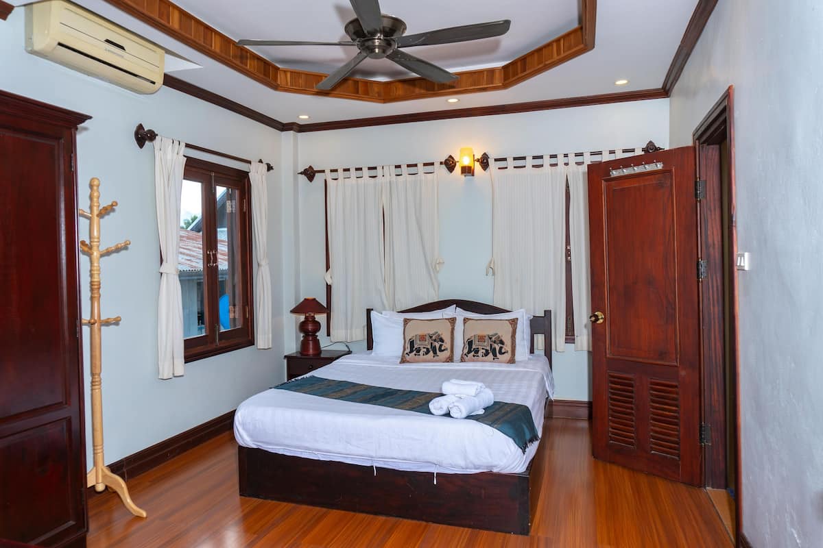 A neatly made bed with white linens and a green throw, a wooden floor, a ceiling fan, and a wooden door.