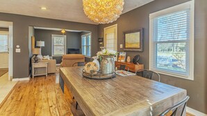 A dining area with a wooden table, chairs, a chandelier, and a view of the outdoors through large windows. - Piccolo Cottage -  Great Location - Walking distance to Douglas Beach! (Douglas)