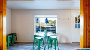 A room with a round glass table and four green stools. There is a window with blue curtains and a view of a backyard. - Beach Cottage Heart of Ocean Drive Sleeps 14 (North Myrtle Beach)