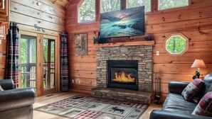 Een gezellig cabin interieur met een stenen openhaard, een televisie erboven en een groot raam met uitzicht op een bos.