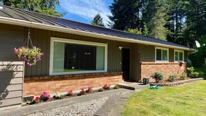 A single-story house with a brick foundation, a metal roof, and a front door. There is a hanging flower basket on the left side of the house, and a small garden with various plants and flowers in front of the door. - Cozy, 3 bedroom home- ideal for couples or families (Forks)