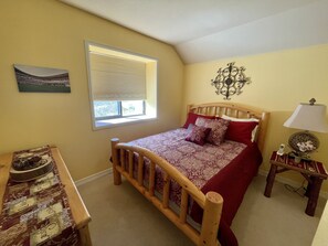 Ein Schlafzimmer mit einem hölzernen Bett, einem Nachttisch mit Lampe, einem Fenster mit Jalousien, einer Wanddekoration und einem kleinen Tisch mit einer Vase.