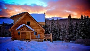 Eine Holzhütte mit schneebedecktem Dach, umgeben von verschneiter Landschaft und einem Wald.