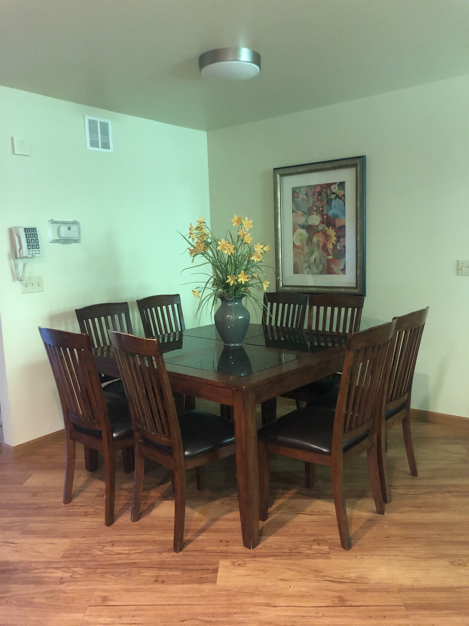 A dining room with a glass-top table surrounded by wooden chairs, a vase with flowers, a ceiling light, and a framed picture on the wall.