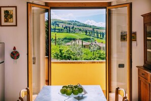 Une pièce avec un balcon donnant sur un paysage verdoyant, une table avec un saladier de pommes et un buffet en bois.