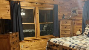 A wooden cabin bedroom with a bed, dresser, and a window with curtains.