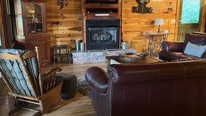 A cozy cabin interior with a wooden floor, a leather sofa, a rocking chair, a fireplace, and a wooden cabinet.