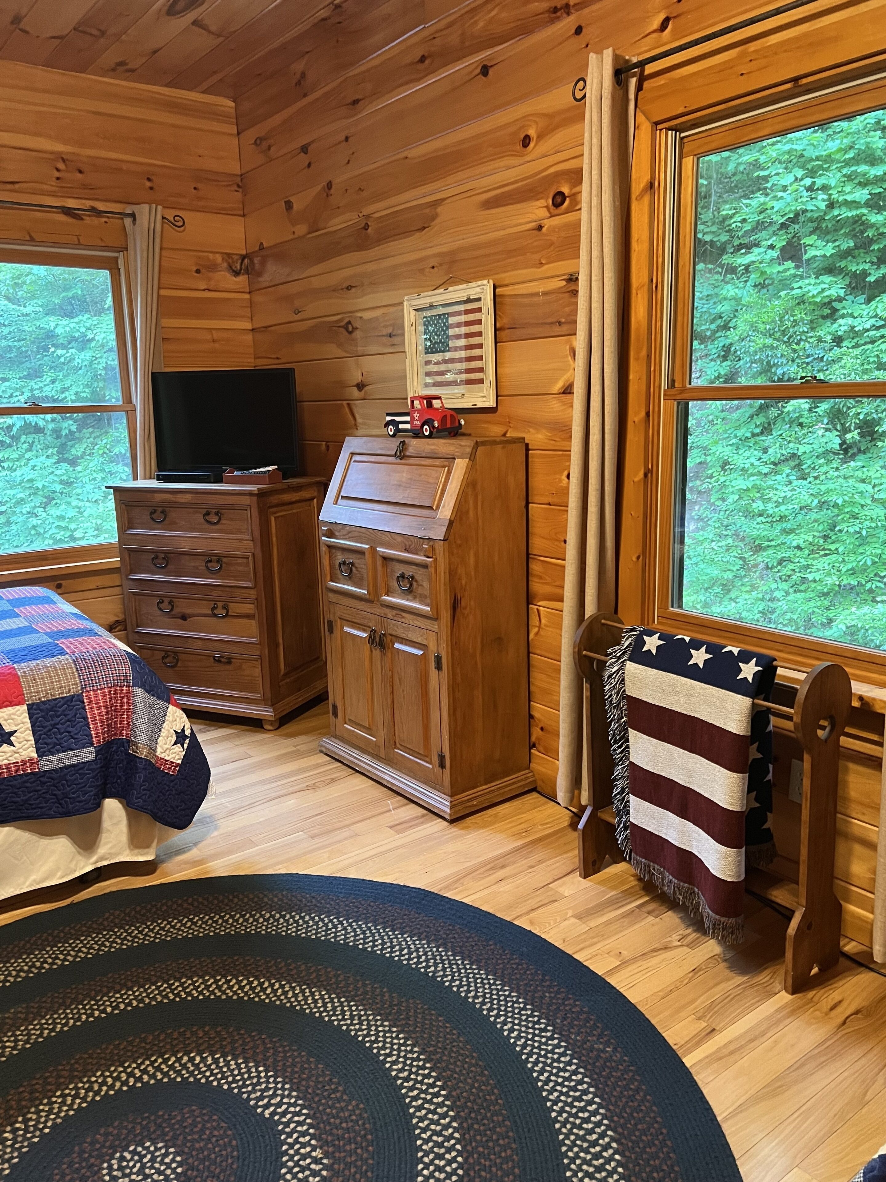 A cozy cabin room with wooden walls, a wooden dresser, a television, a quilt, and a view of a forest through large windows.