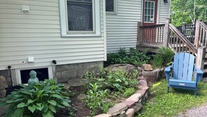 A house with a front porch, a blue Adirondack chair, and a garden with plants and a stone border. - Village Hideaway, 1 bdrm efficiency, private entrance. On the Rail Trail. (Saranac Lake)