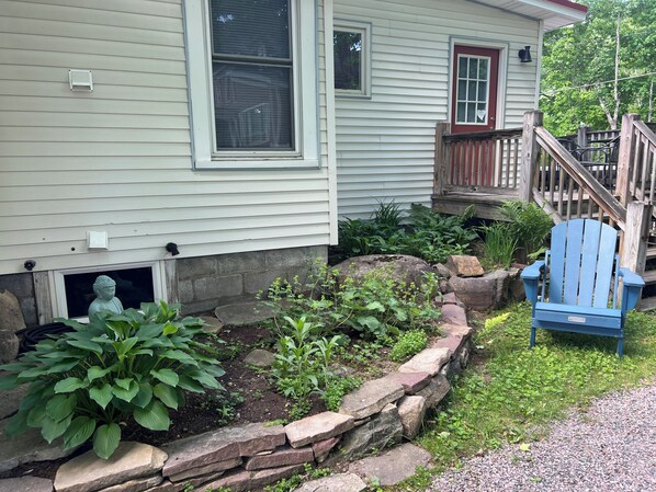 Una casa con porche, una silla Adirondack azul y un jardín con plantas y un borde de piedra.