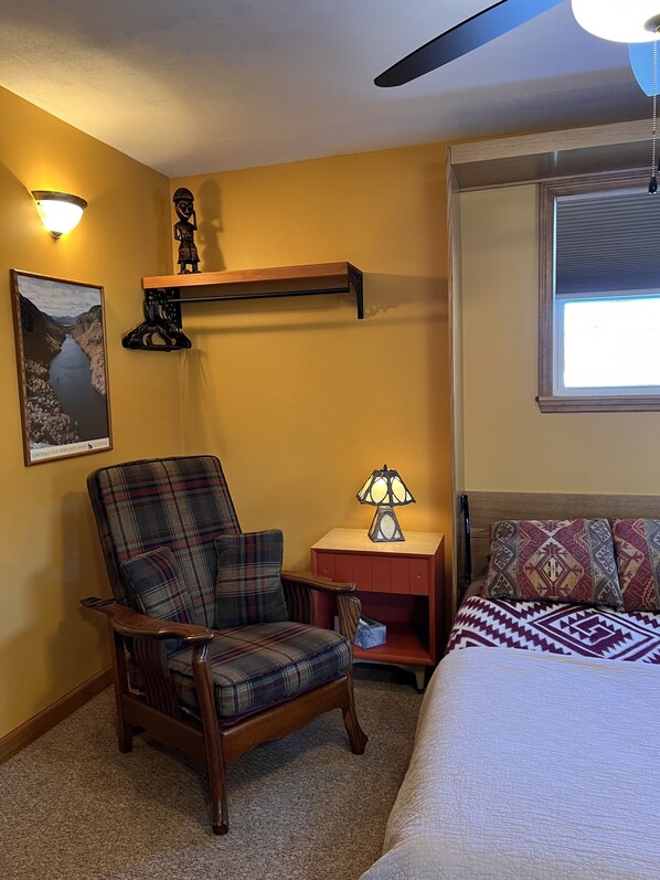 A bedroom with a plaid armchair, a bedside table with a lamp, a bed with patterned pillows, a wall-mounted shelf with a statue, and a framed picture on the wall.