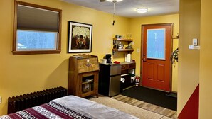 A compact room with a ceiling fan, a window with blinds, a framed picture, a small wooden cabinet, a refrigerator, and a door with a peephole. - Village Hideaway, 1 bdrm efficiency, private entrance. On the Rail Trail. (Saranac Lake)