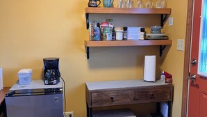 A compact kitchenette with a refrigerator, coffee maker, and a microwave. There are shelves above holding kitchenware and a small table with a drawer. - Village Hideaway, 1 bdrm efficiency, private entrance. On the Rail Trail. (Saranac Lake)