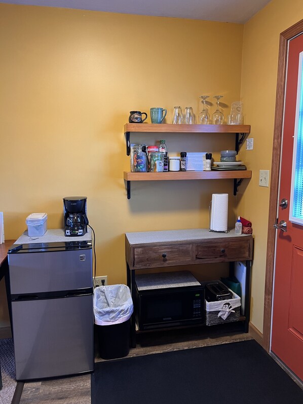 Una kitchenette compacta con refrigerador, cafetera y microondas. Hay repisas con utensilios de cocina y una mesita con un cajón.