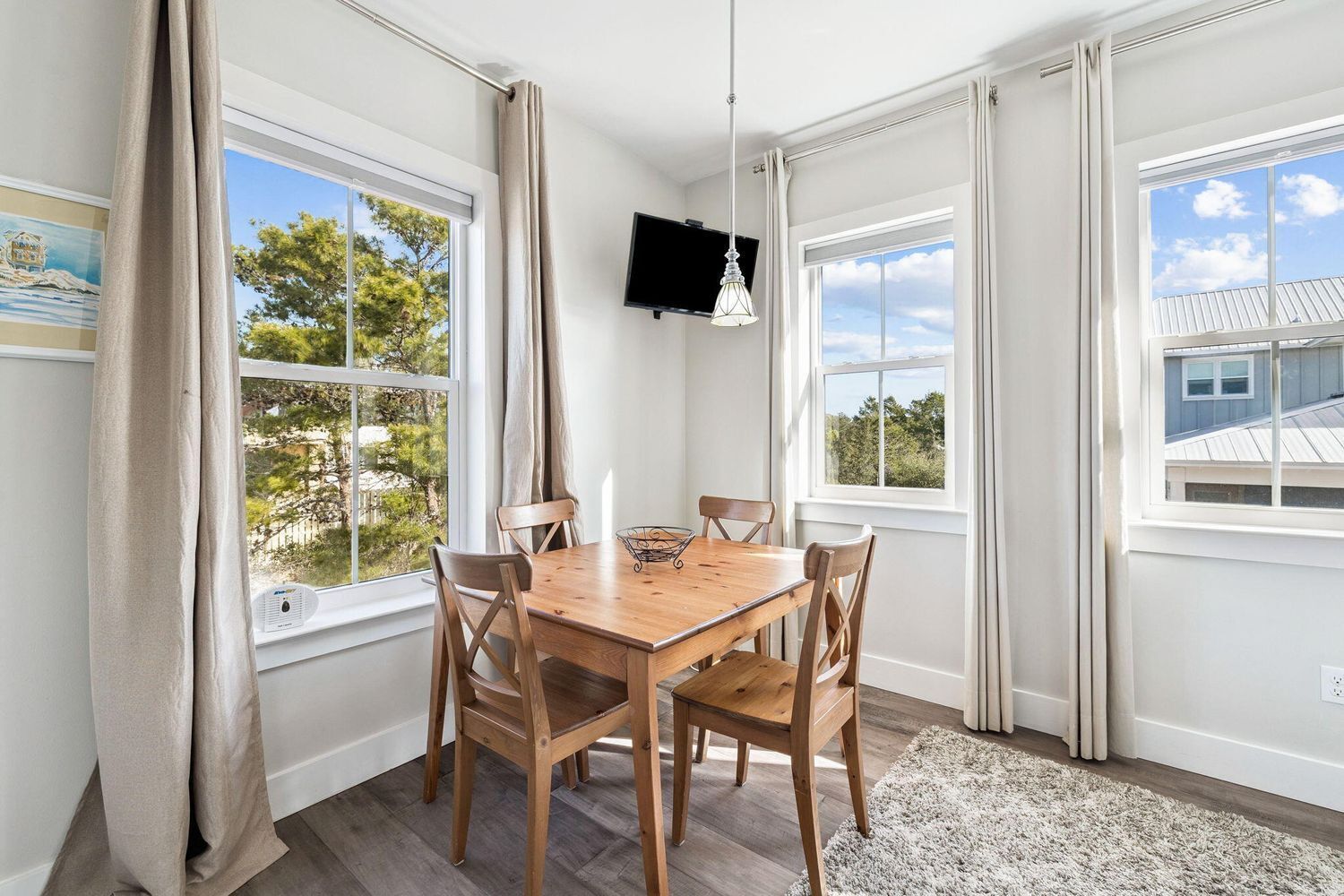 Un área de comedor con una mesa y sillas de madera, ventanas grandes con cortinas, un televisor de pantalla plana y una lámpara de techo.