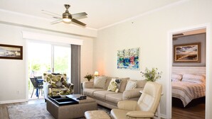A living room with a sofa, armchairs, a coffee table, and a ceiling fan. There is a door leading to a bedroom with a bed.