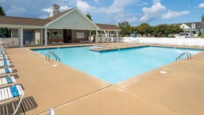 A rectangular swimming pool with a clear blue water surface, surrounded by a beige deck. There are lounge chairs aligned along the pool's edge, and a covered area with a sloped roof is visible in the background.