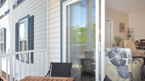 A covered porch with a wooden table and chairs, a sliding glass door leading to an interior living space with a sofa and a chair, and a decorative flower on the wall.
