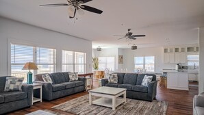 Un espacio habitable amplio con dos sofás grises, una mesa de centro y una mesa de comedor. Se observan ventiladores de techo, ventanas grandes y una zona de cocina.