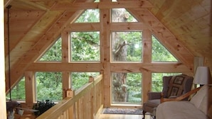 Une cabane en bois avec une chambre à l’étage, comprenant un lit, une chaise et un bureau. De grandes fenêtres offrent une vue sur la forêt environnante.