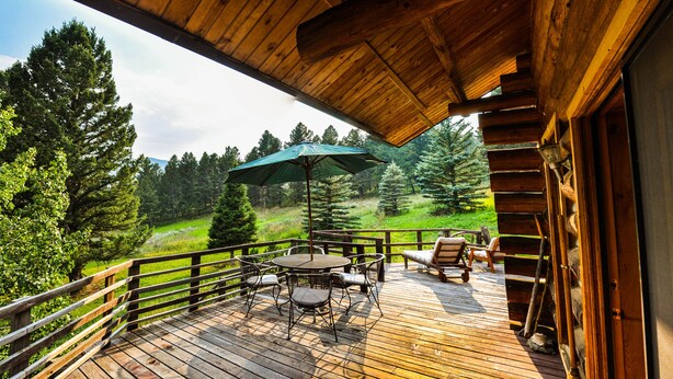 A wooden deck with outdoor furniture and a green umbrella, overlooking a forested area. - Mountain Log Home Close to Historic Downtown Bozeman & Bridger Bowl. (Bozeman)