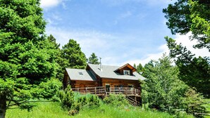 A log cabin with a wooden deck and a small porch, surrounded by lush greenery and trees. - Mountain Log Home Close to Historic Downtown Bozeman & Bridger Bowl. (Bozeman)