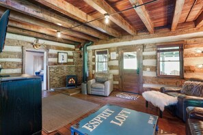 A rustic log cabin interior with exposed wooden beams, a fireplace, leather sofas, and a large window.