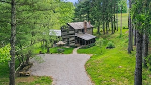A rustic log cabin with a gravel driveway, surrounded by dense forest. - ONE OF A KIND HISTORIC LOG CABIN ON 10+ ACRES IN HOCKING HILLS (Sugar Grove)