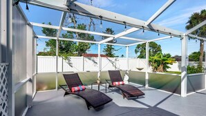 A covered outdoor area with two lounge chairs and a small table, surrounded by a white fence and greenery. - Paradise Palace and POOL home (Cape Coral)
