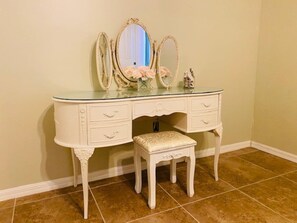 A white vanity with three mirrors, a bench, and a vase with flowers.