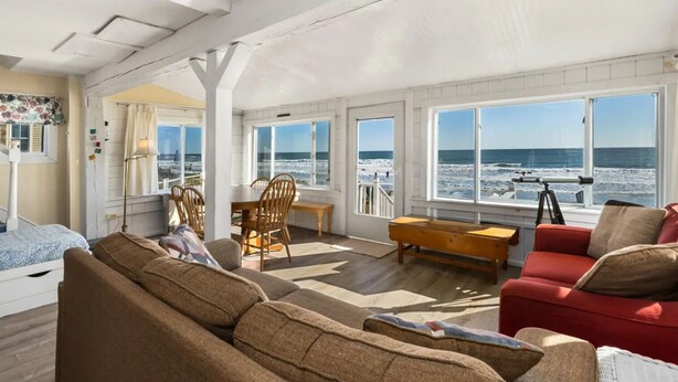 A spacious room with a large window overlooking the beach, a wooden dining table, and a red sofa. - Enjoy sunrises on the Private Beach - 4 bedrooms (Wells)