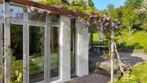 Ett hus med stora glasdörrar, en träpergola med blommande klätterväxter och en trädgårdsdel med ett bord och stolar.