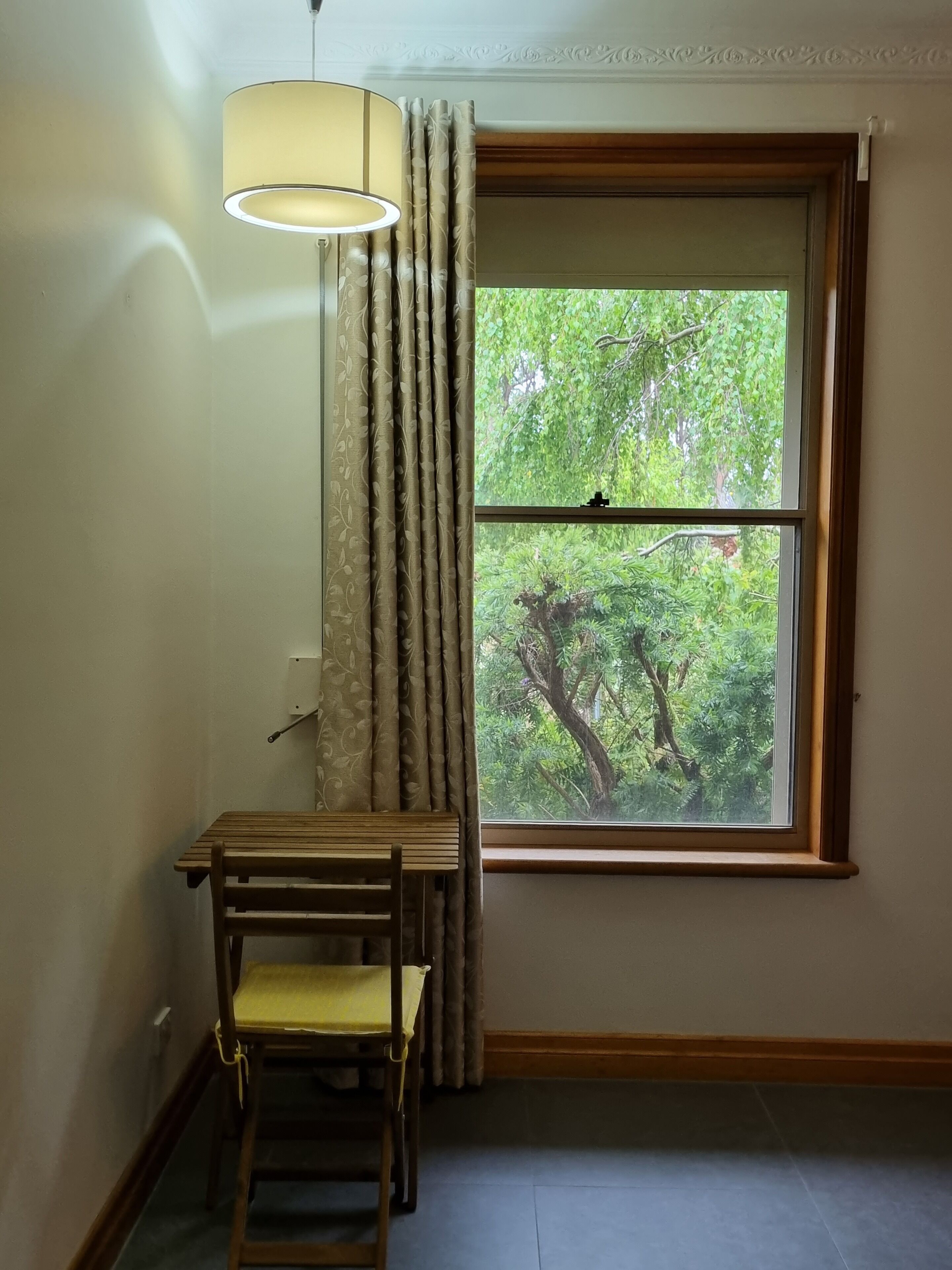 Une pièce avec une fenêtre, un bureau en bois, une chaise et une lampe suspendue.