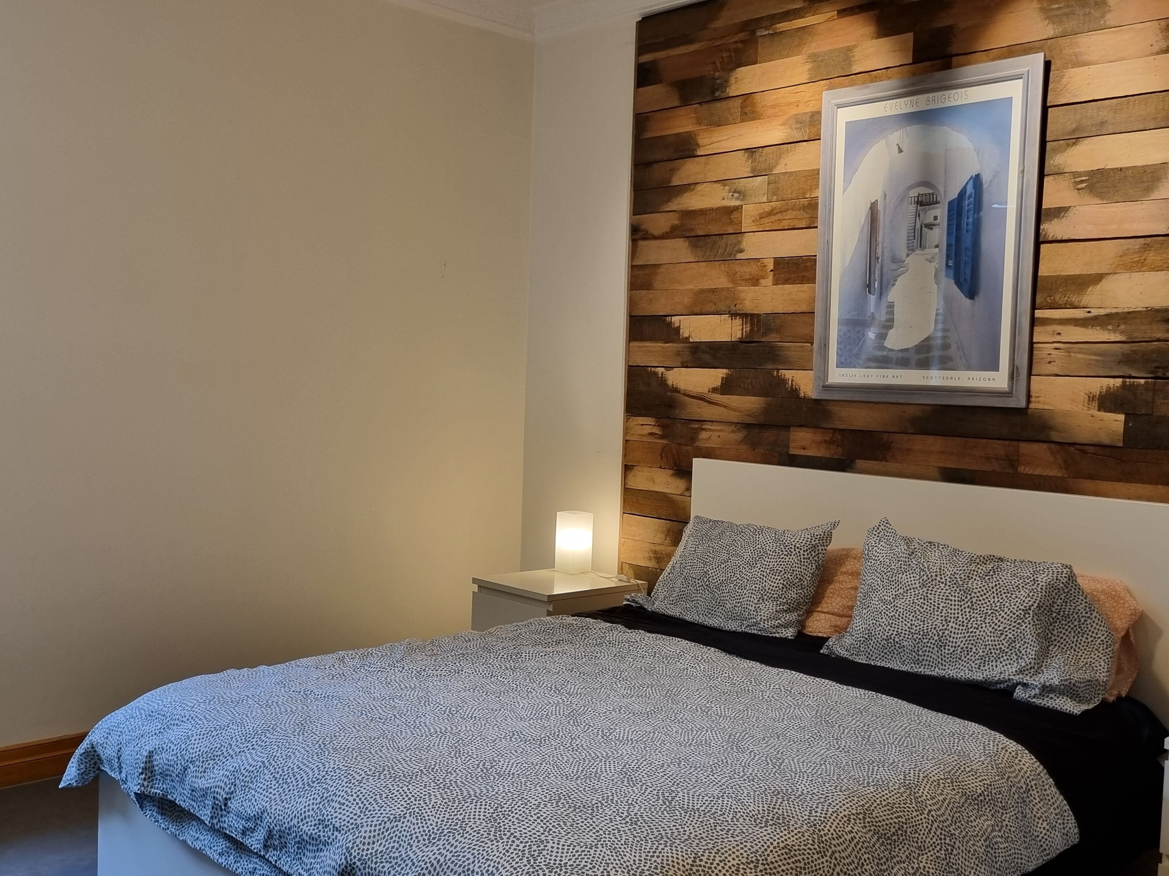 Un dormitorio con cama, dos almohadas, una mesita de noche, un cuadro colgado en la pared y un panel de madera.
