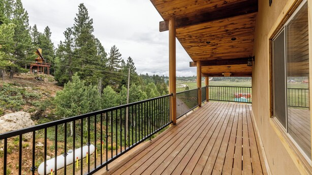 Eine Holzterrasse mit schwarzer Reling, mit Blick auf eine bewaldete Gegend und ein weiter entfernes Gebäude.