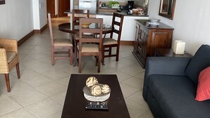 Una sala de estar con un sofá oscuro, una mesa de centro con un bol de pan y un comedor con sillas de madera, con una cocina al fondo.