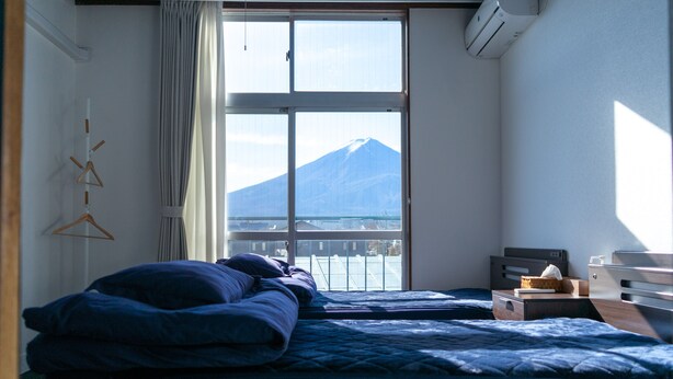 A bedroom with two beds, a window with a view of a mountain, and a bedside table with a lamp. - FUJI GUESTHOUSE AO - Hostel (Fujikawaguchiko)