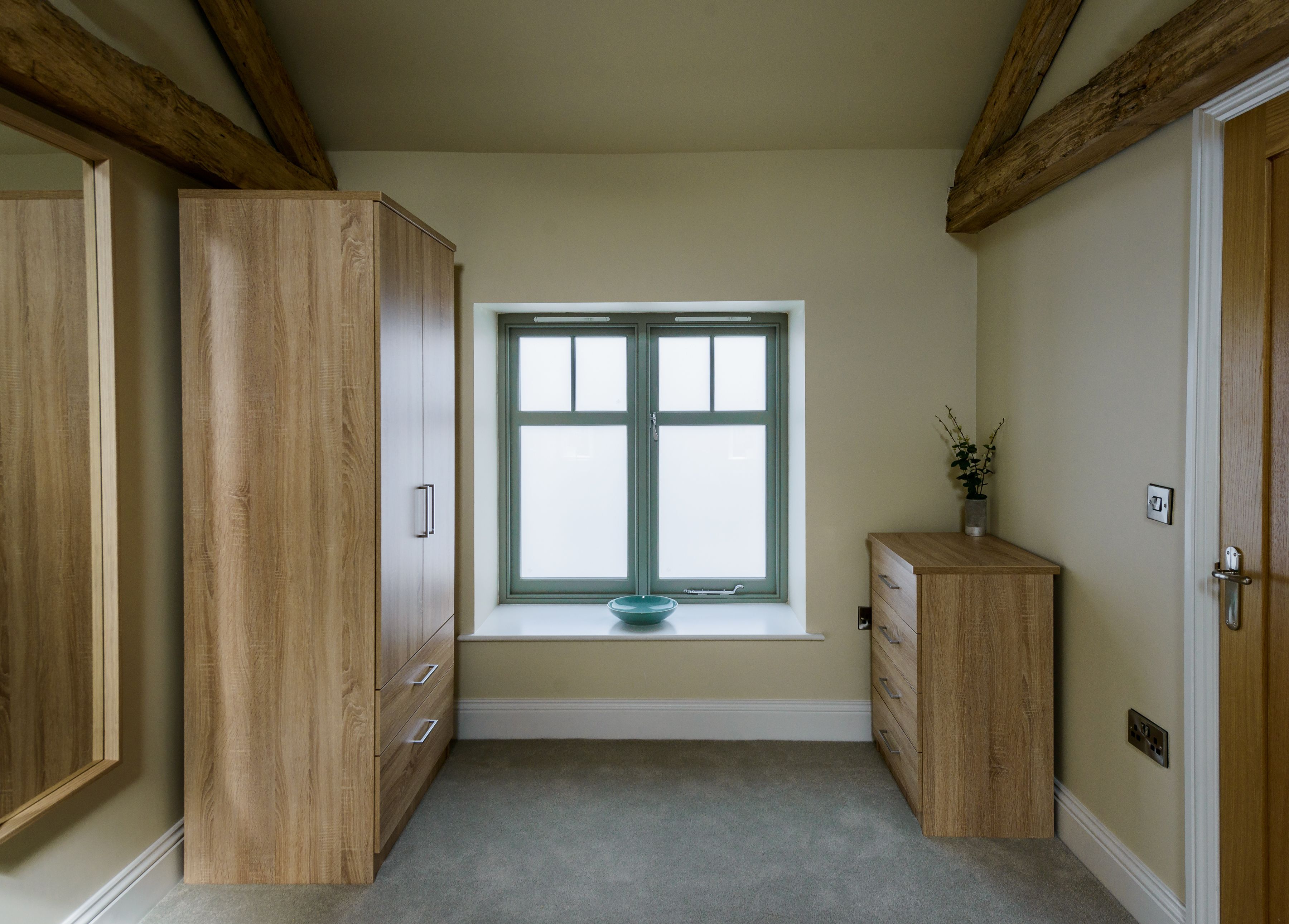 A room with a large wooden wardrobe, a small window with a bowl on the sill, and a wooden cabinet.
