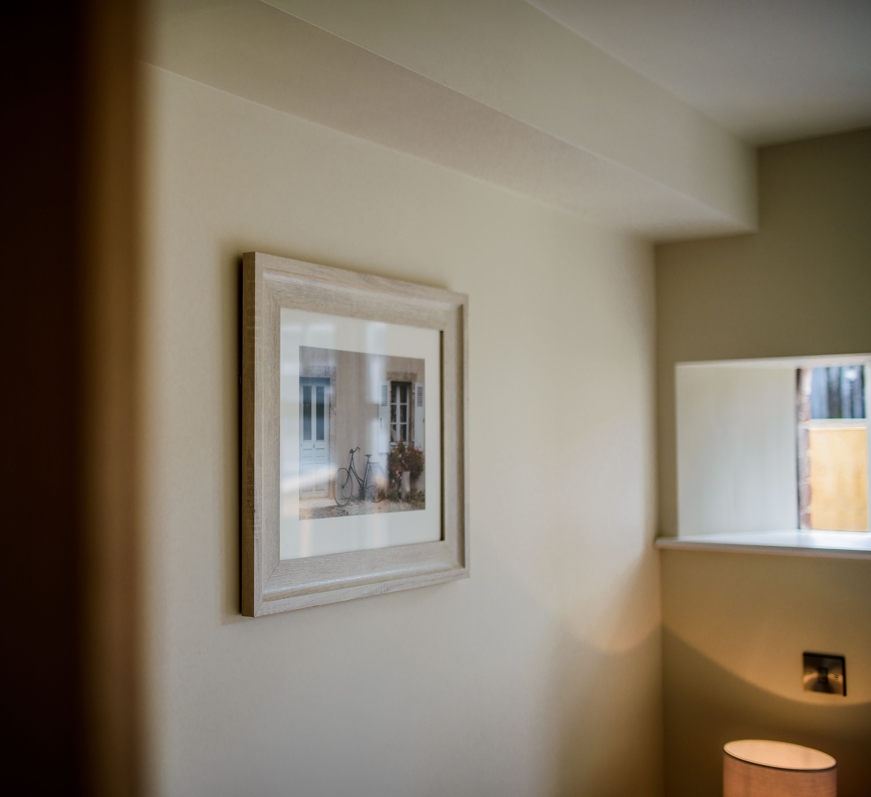 A framed picture on a wall, a lamp, and a window with a view of a building.