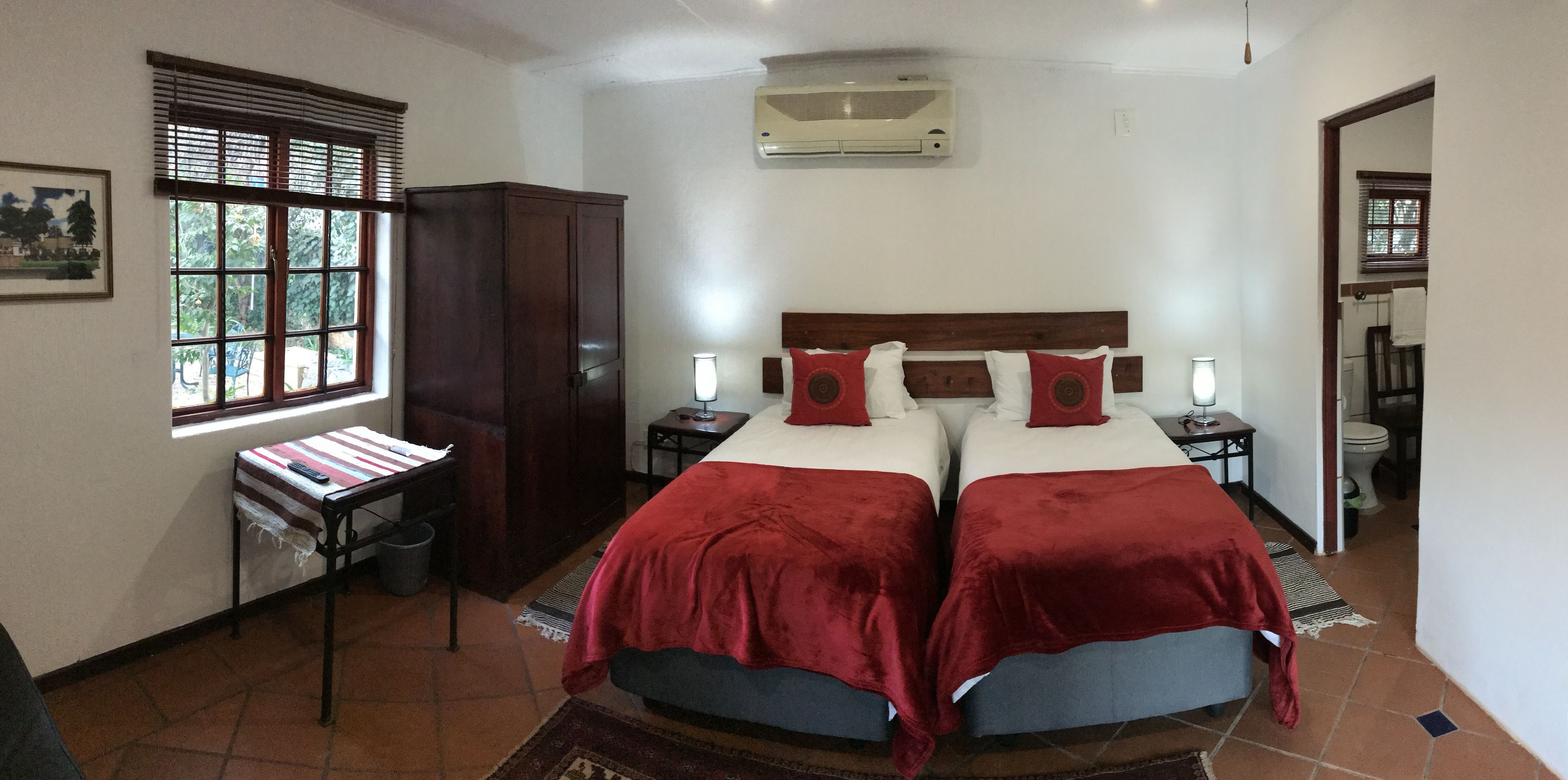 A bedroom with two beds, a wardrobe, a desk with a lamp, and a window with blinds.