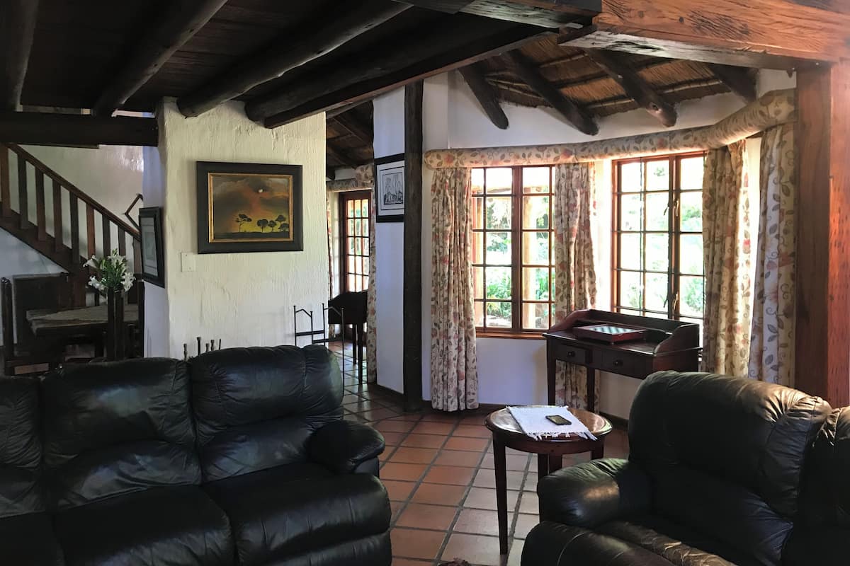 A cozy living room with a high ceiling, wooden beams, a staircase, and a view through large windows with curtains.