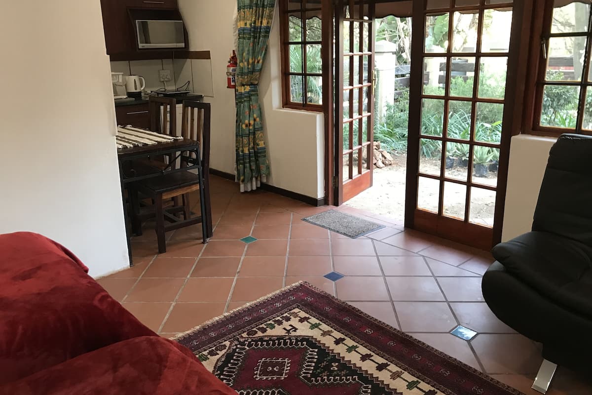 A room with a kitchen area, a dining table with chairs, a large window with a view of the outdoors, and a red sofa.