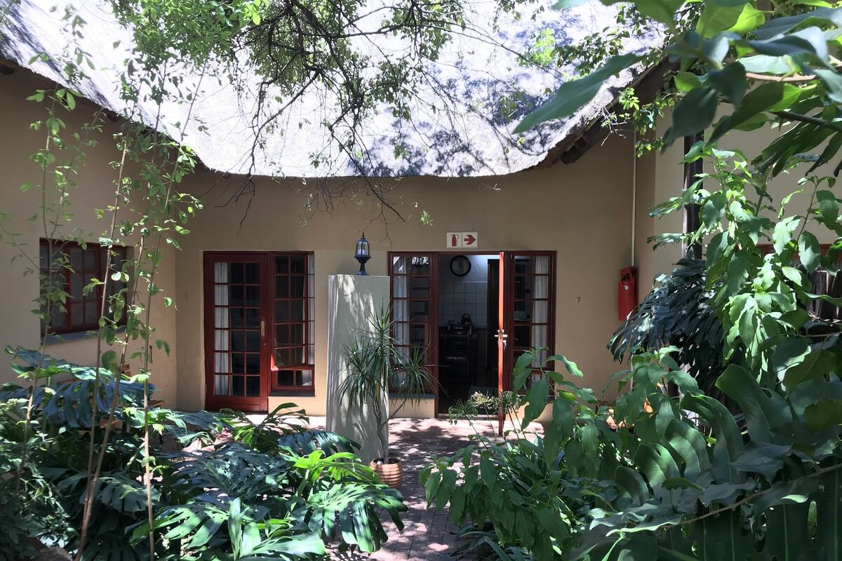 A courtyard with a thatched roof, potted plants, and a door leading to a room.