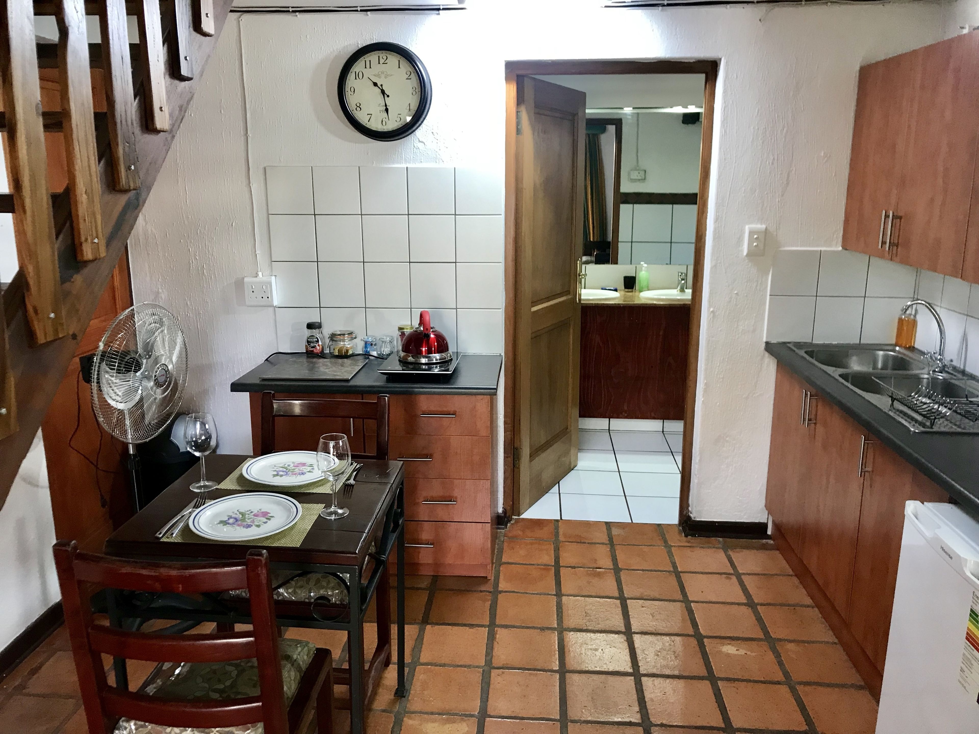 A compact kitchen with a dining table set for two, a clock on the wall, and a fan near the table.