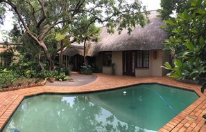 A pool area with a thatched-roof building, surrounded by lush greenery and potted plants. - Greenwoods Self-Catering (Pretoria)