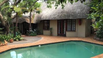 A thatched-roof house with a swimming pool, surrounded by lush greenery and potted plants.