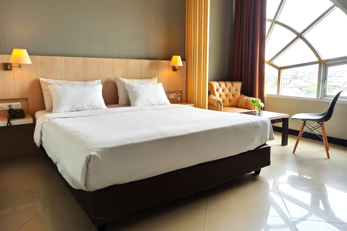 Quarto de hotel com uma cama grande, duas luminárias de cabeceira, uma janela com cortinas, uma pequena mesa com uma planta e uma cadeira.