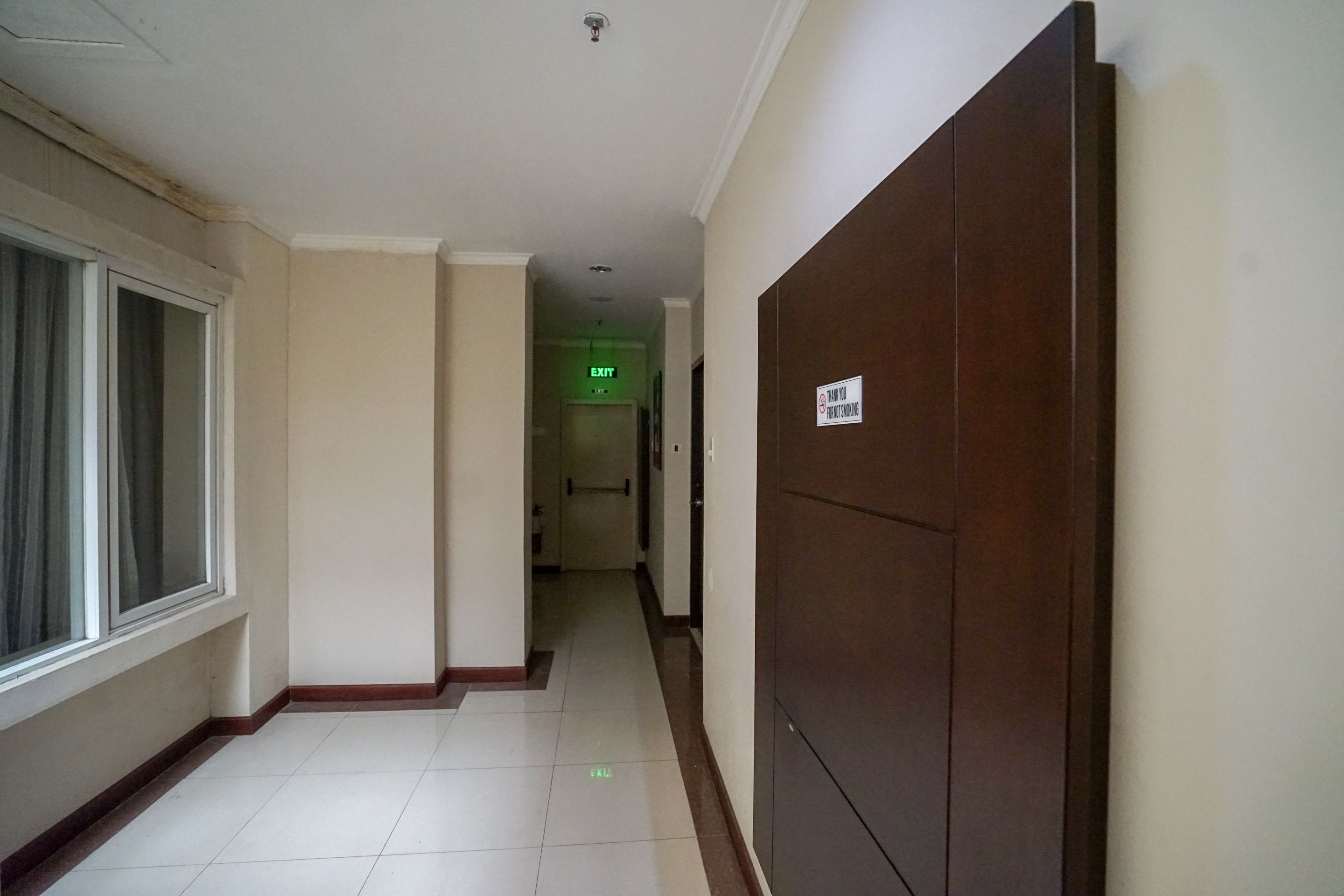 A hallway with a wooden door on the right, a window on the left, and an exit sign in the distance.